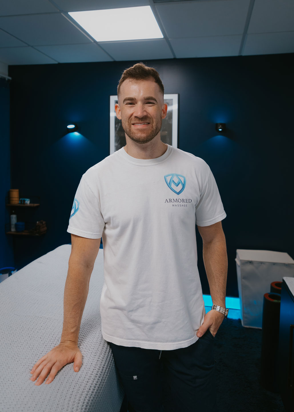Stephen Preyer smiling at the camera sitting behind his massage table, he looks friendly and cheerful, his hands are resting on the massage table, and behind him is a wall with an image of a knight behind him with a cape.