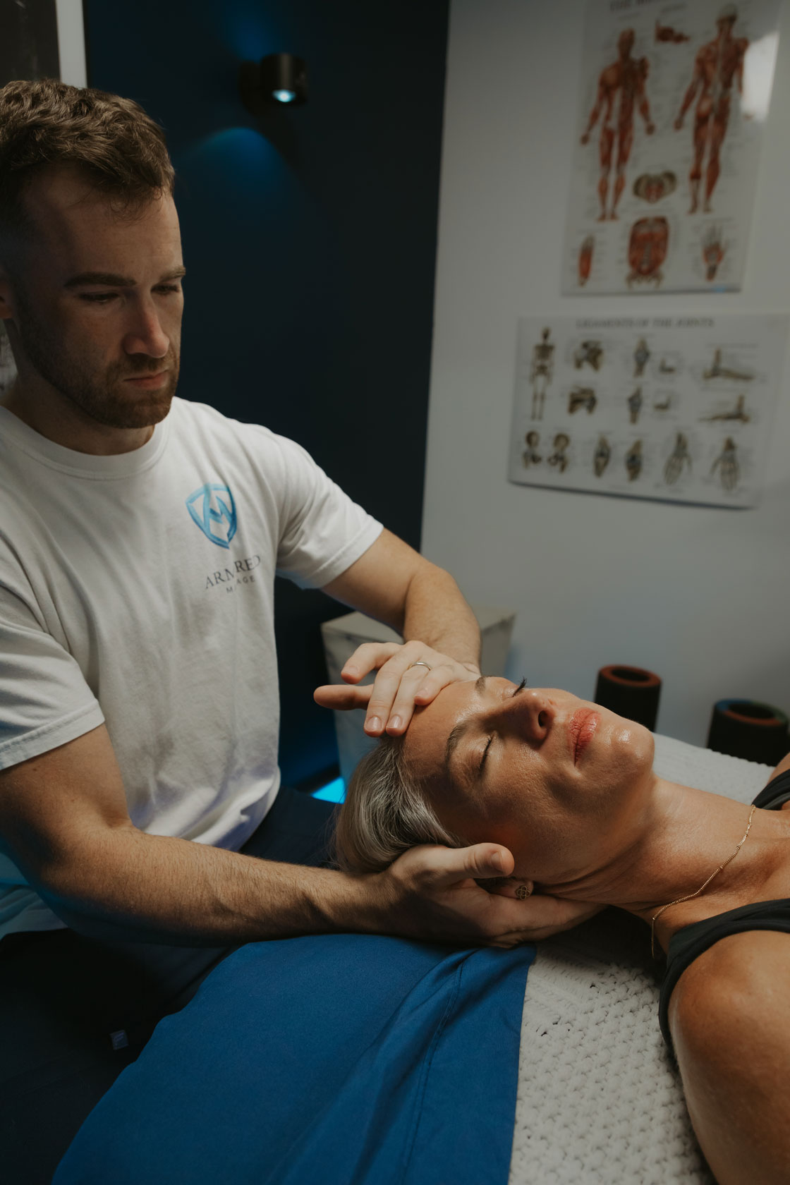 Therapist wearing a white shirt gently stretching a woman's neck while she lies on a massage table covered with a blue blanket in a therapy room with anatomy posters.
