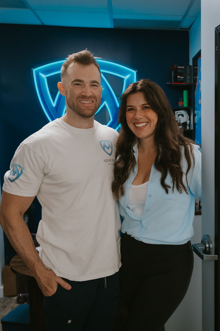 Smiling man in a white logo t-shirt with his arm around a smiling woman in a light blue shirt standing in an office with a blue illuminated logo on the dark wall behind them.