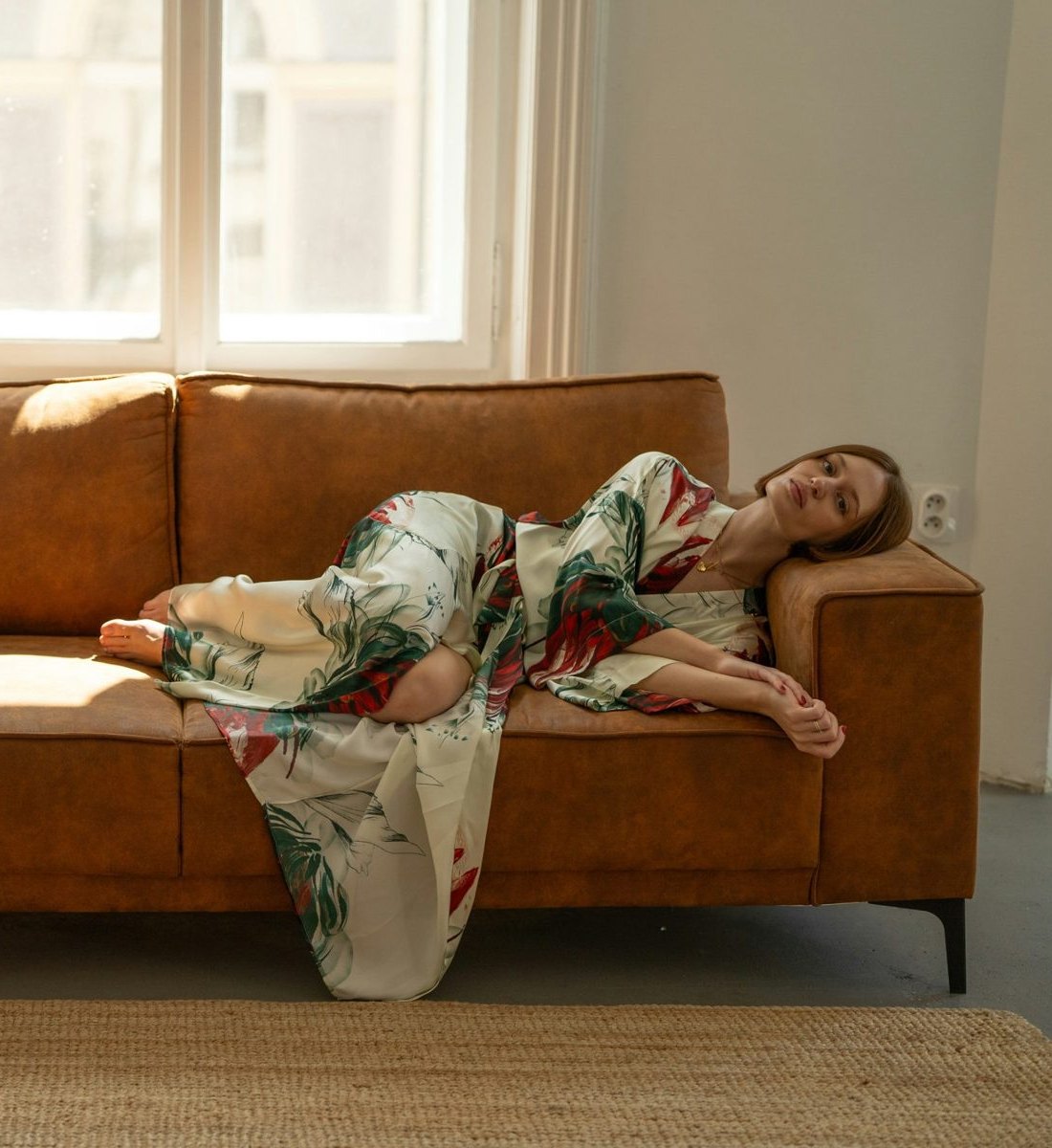 Woman reclining on a tan leather sofa in a cream silk kimono with painted red and green botanicals