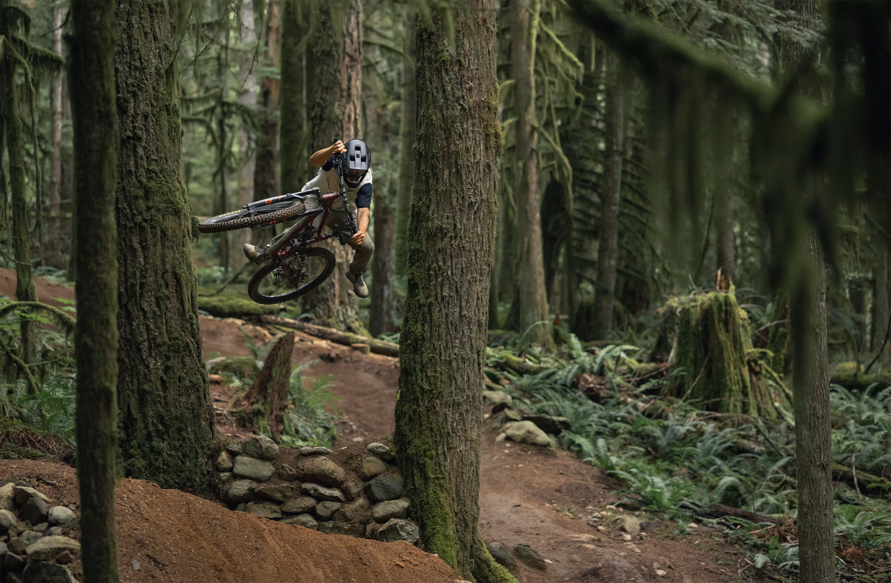 Mountain biker performing a jump trick on a dirt trail in a dense forest.