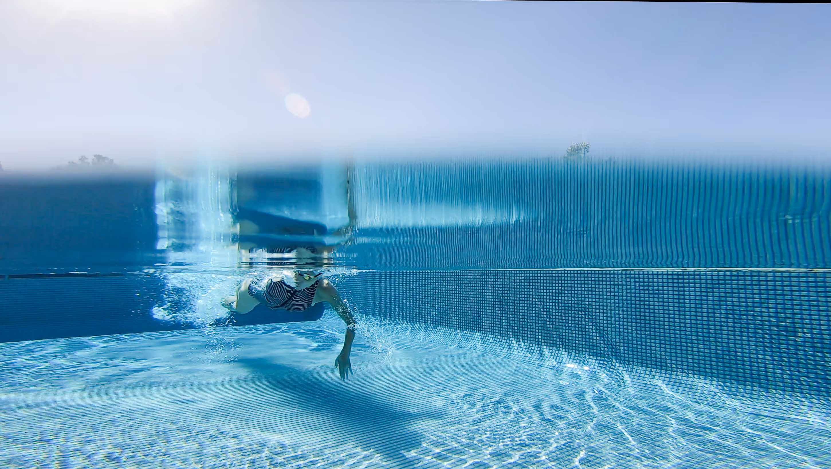 Person swimming underwater in a clear blue pool with sunlight filtering through.