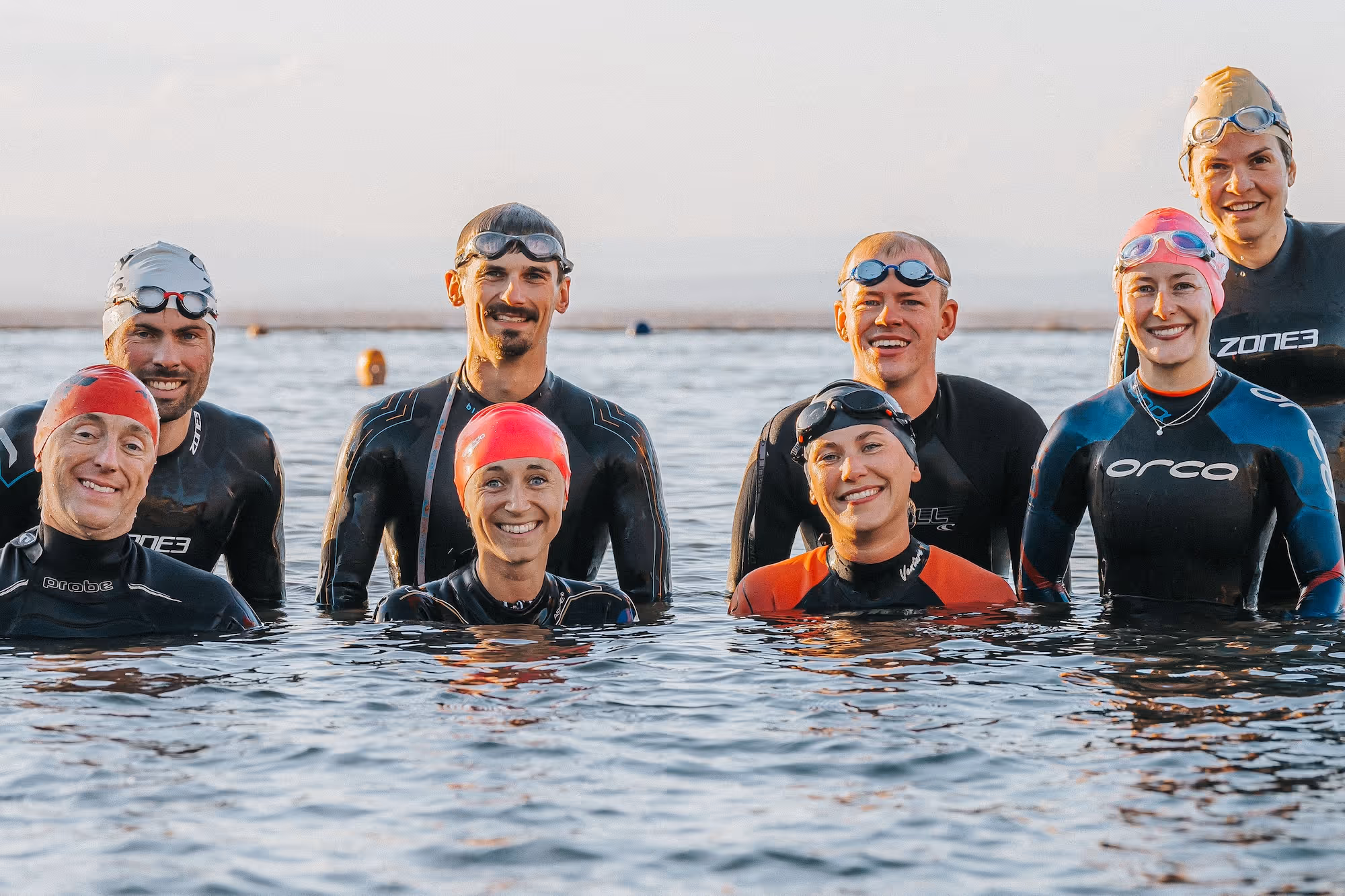 Group of seven swimmers wearing wetsuits, swim caps, and goggles standing waist-deep in calm water, smiling toward the camera at sunset.