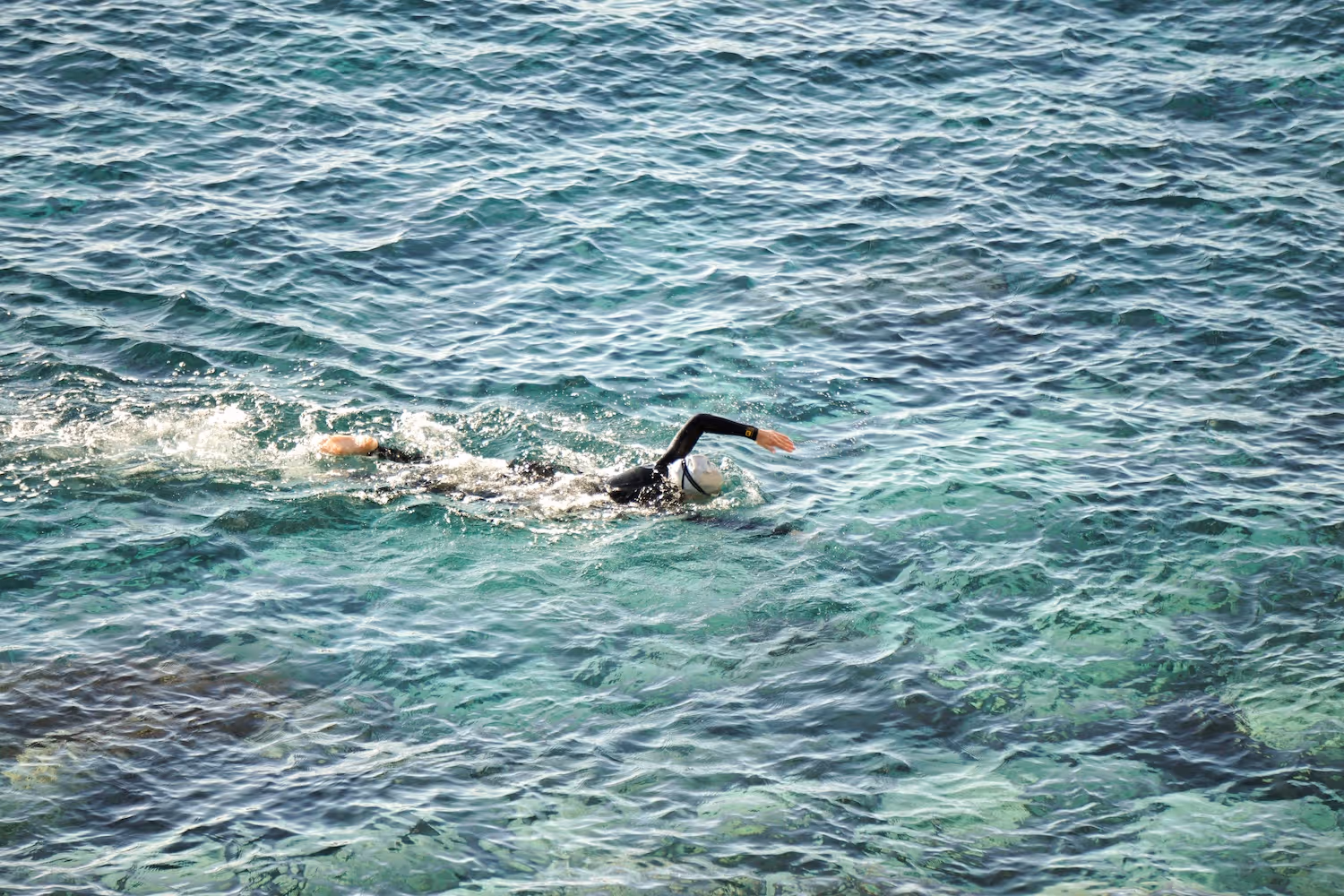 Swimmer wearing a wetsuit and swim cap swimming freestyle in clear blue ocean water.