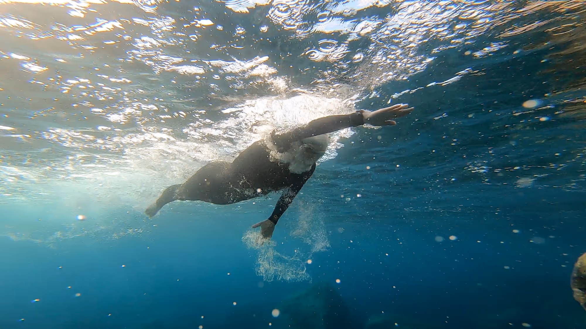 Person swimming underwater wearing a black wetsuit, making a freestyle stroke with bubbles around.