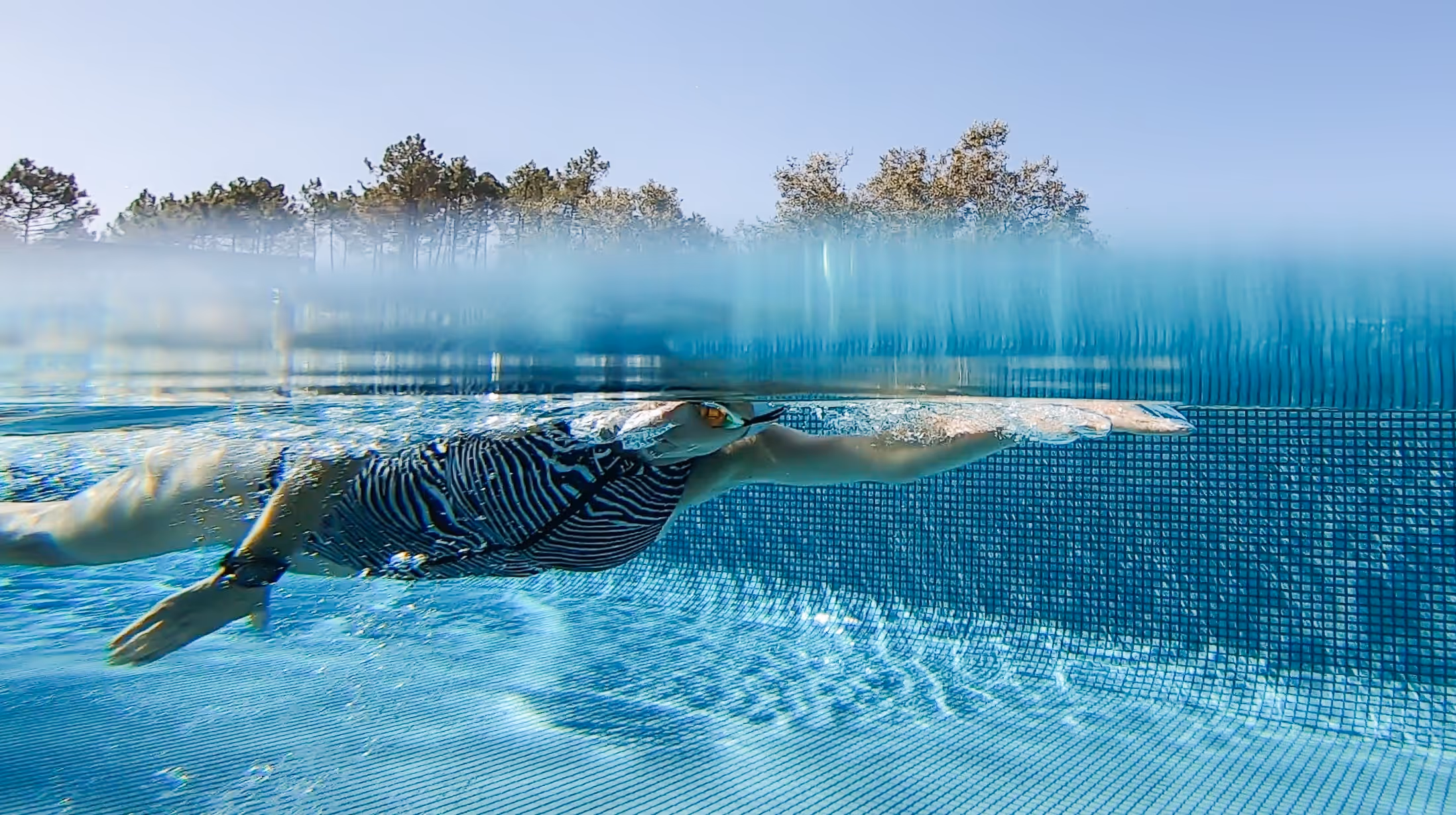 Swimmer in striped swimsuit glides underwater near the edge of a clear blue pool with trees visible above water.