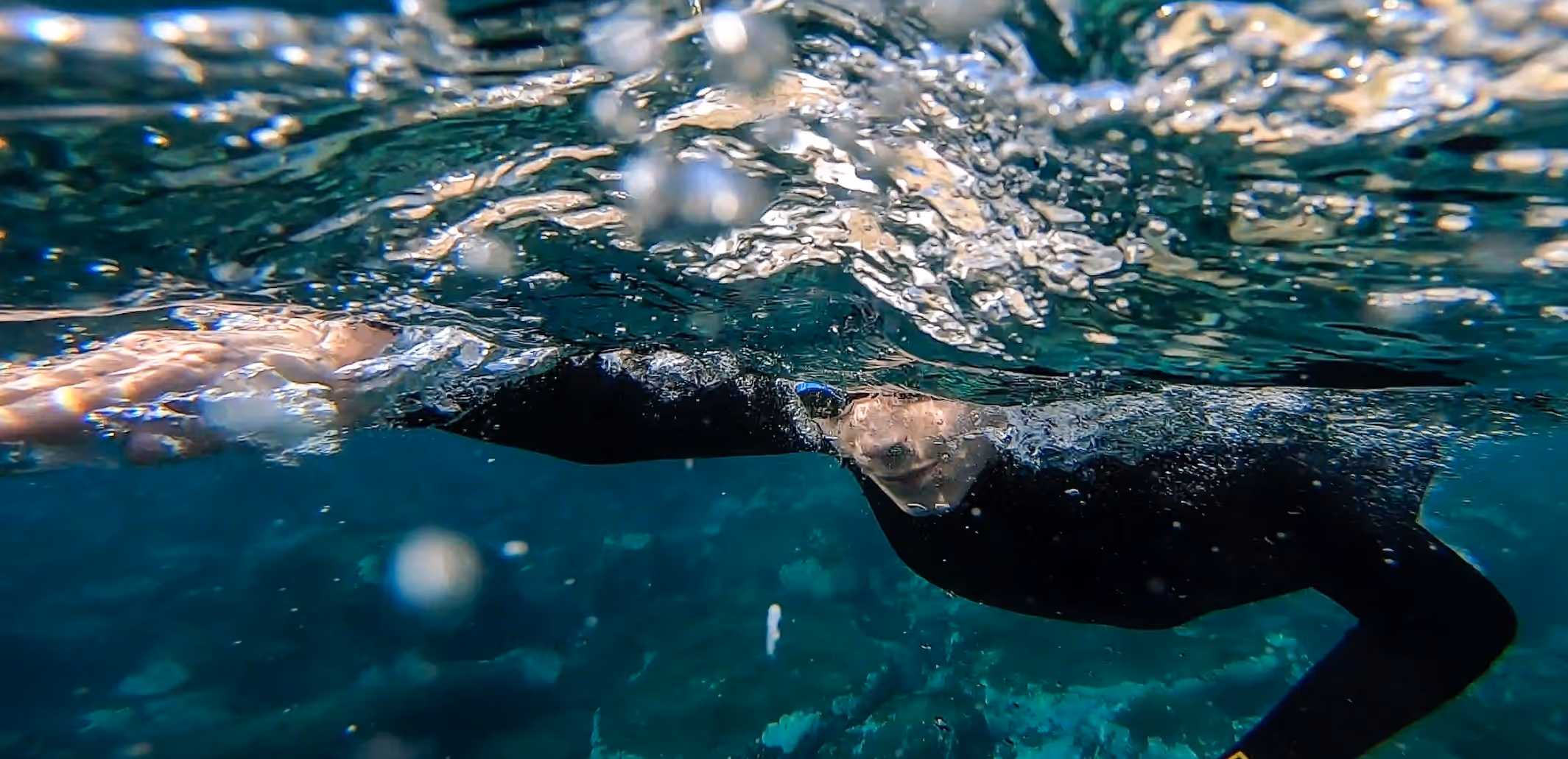 Underwater view of a swimmer in a black wetsuit extending one arm forward through clear blue water.