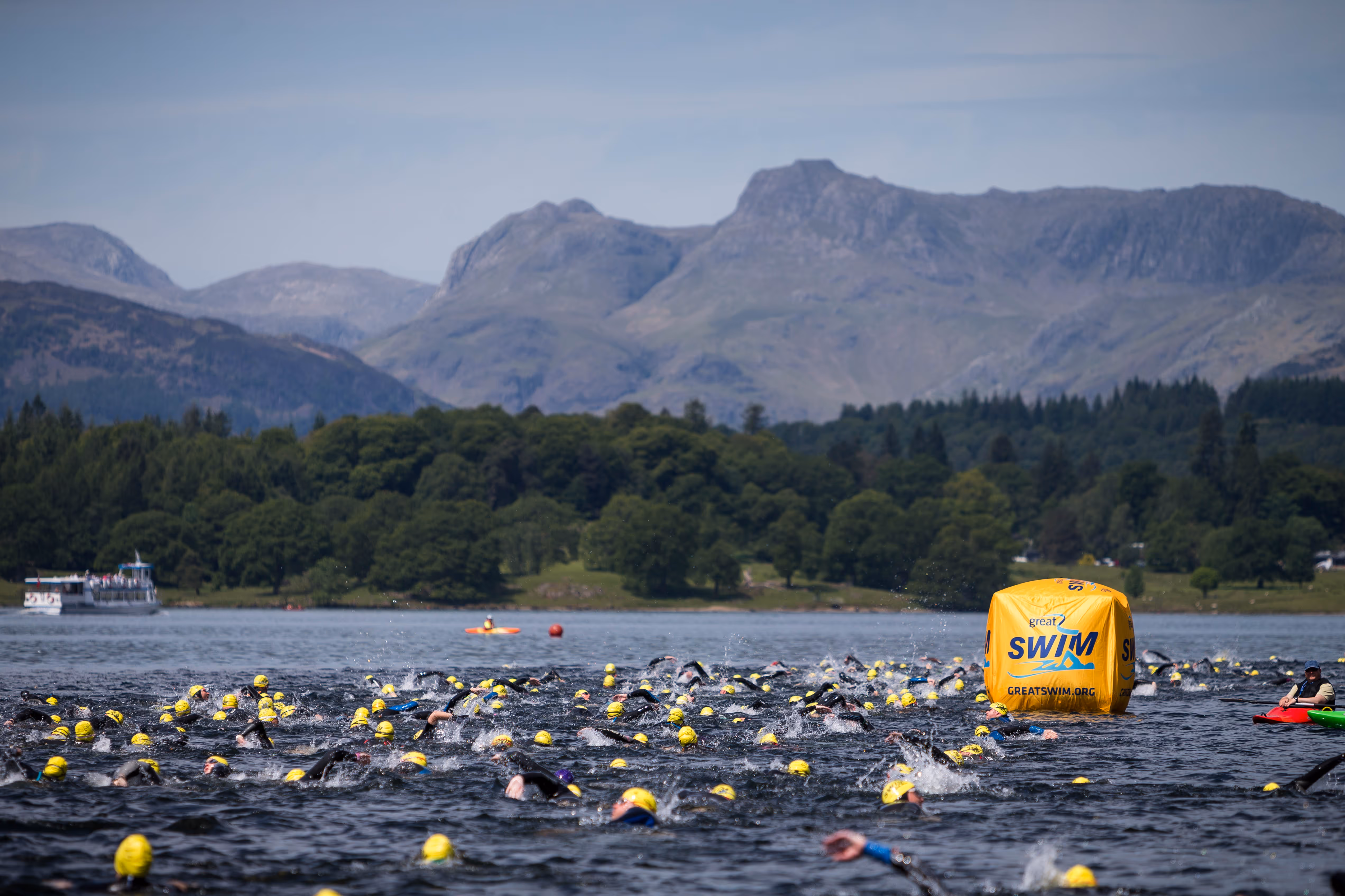 Large group of swimmers wearing yellow caps in open water near a yellow race buoy marked Great Swim, with mountains and forest in the background.