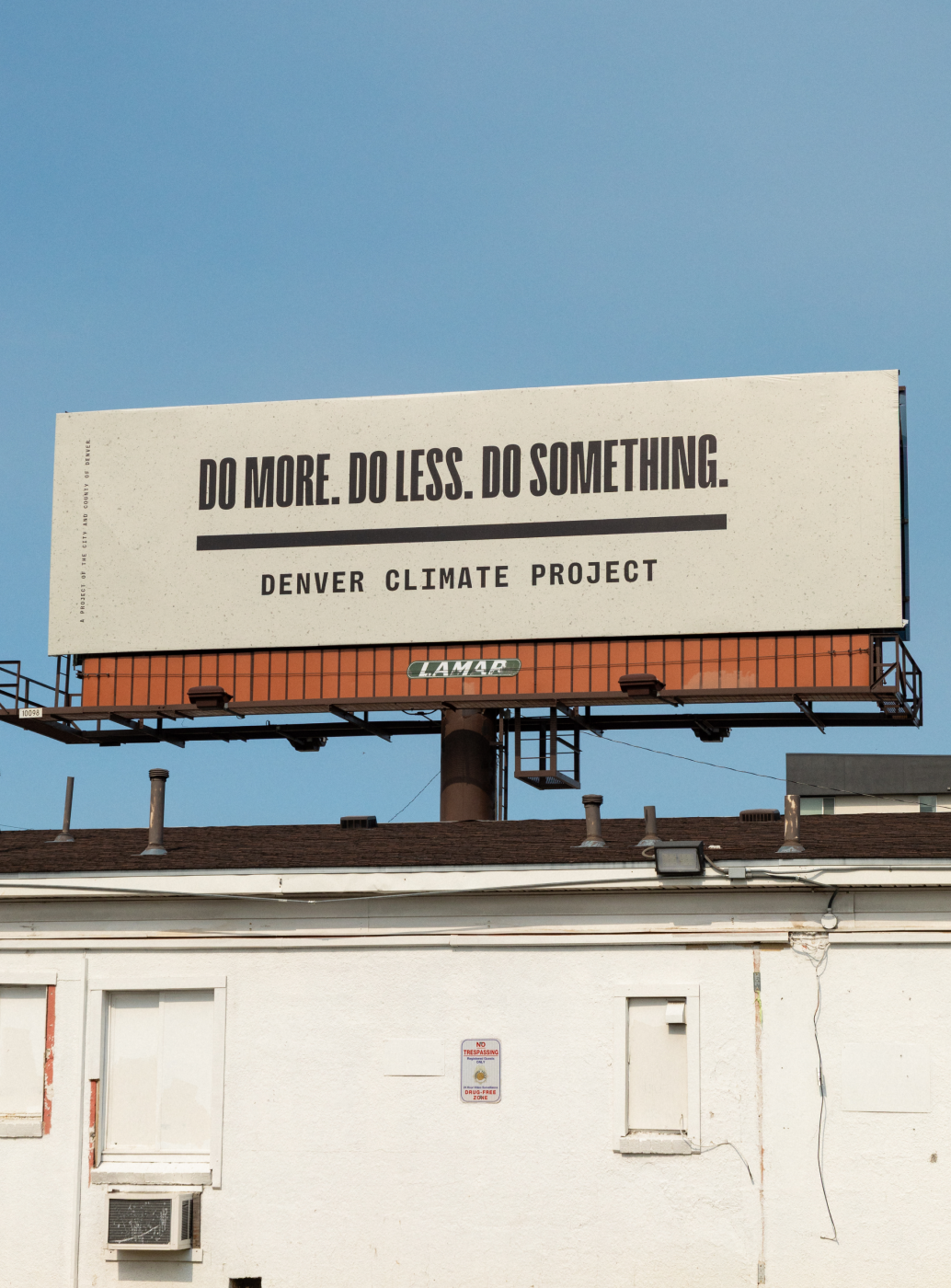 Photo of a tan billboard on a sunny day that reads "DO MORE. DO LESS. DO SOMETHING. Denver Climate Project" in black all caps type in front of a blue sky.