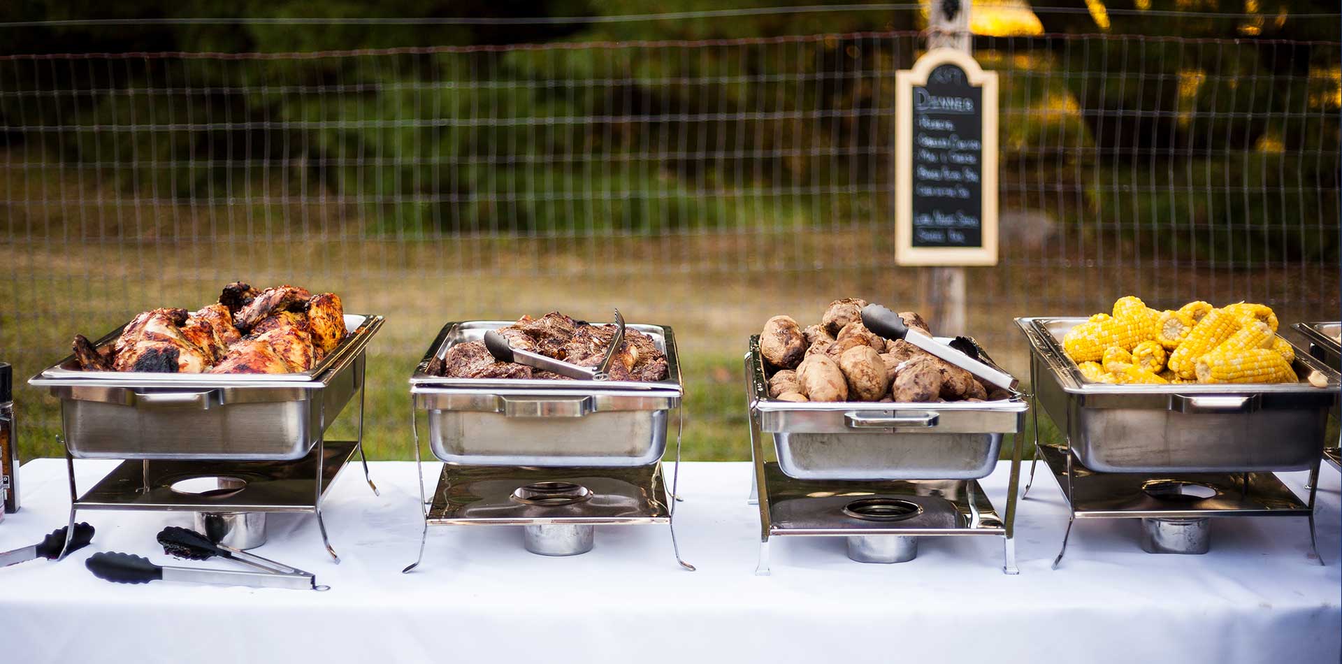 Food warmers on a table with barbecued steak, chicken, potatoes and corn in each warmer