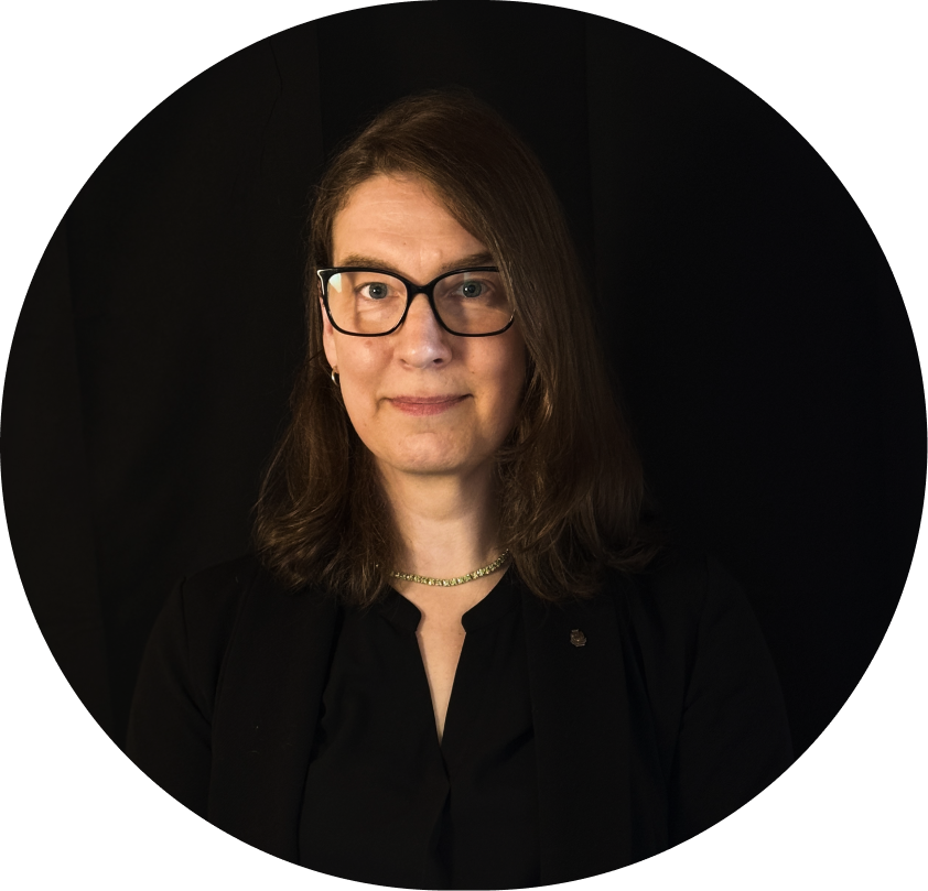Formal portrait of Tana Green, looking directly forward. All black background and clothes. Shoulder length brown hair, black frame glasses, green crystal necklace, American Institute of Architects pin, simple smile.