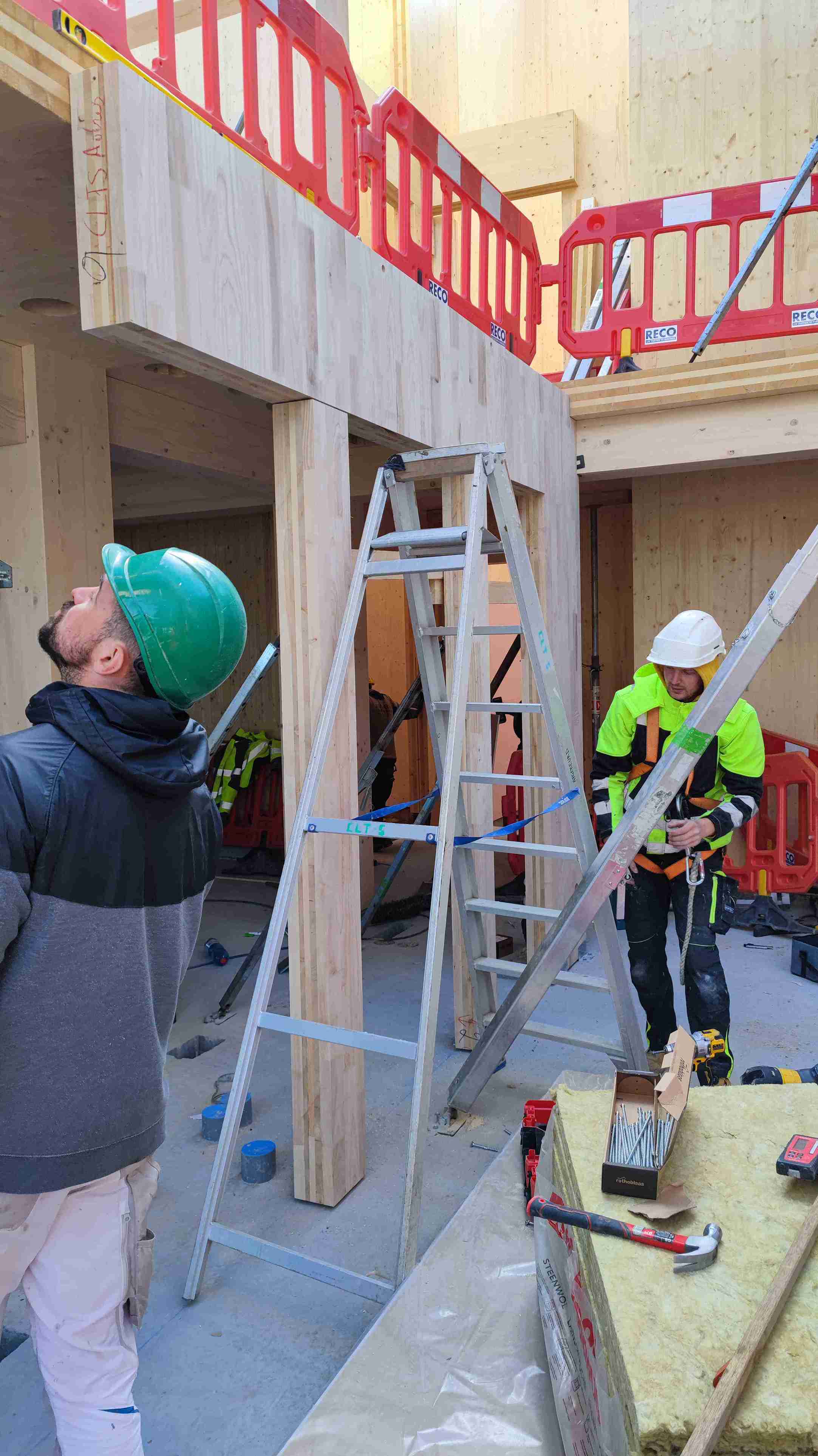 In de ruwbouw zijn C-CLT-kolommen en liggers zichtbaar terwijl vakmensen de houten elementen monteren, omringd door ladders, isolatiemateriaal en gereedschap.