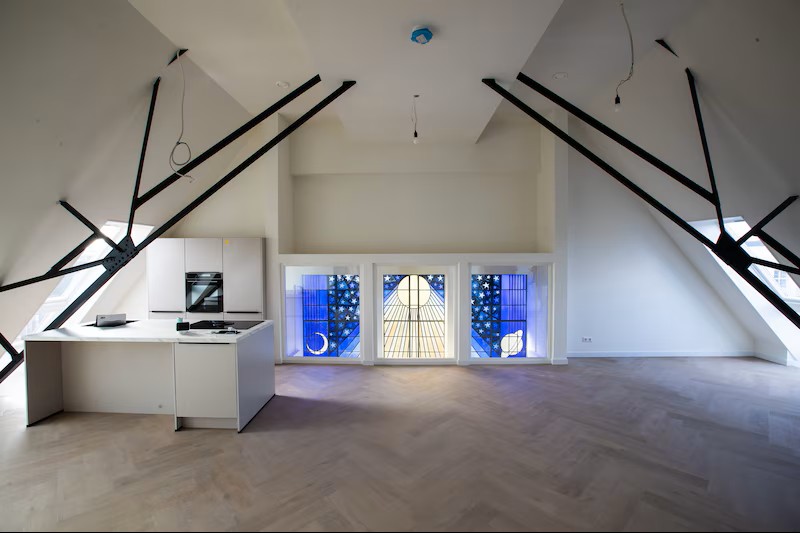 Licht en open penthouse onder een schuine kap met zichtbare zwarte dakconstructie, houten vloer en minimalistische keuken. Grote glas-in-lood ramen brengen gekleurd daglicht binnen en geven de ruimte karakter en hoogte.