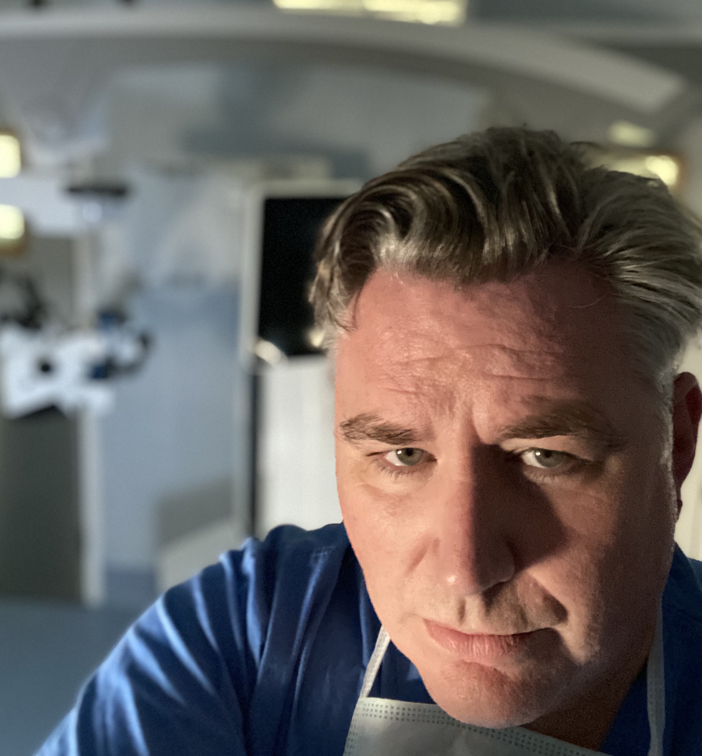 Close-up of a middle-aged man with gray-streaked hair wearing a blue shirt and surgical mask under his chin in a medical or clinical setting.