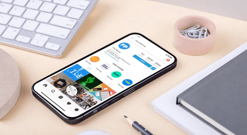 A smartphone with social media app icons displayed on the screen, placed on a desk beside a keyboard, an earphone case, and a notebook.