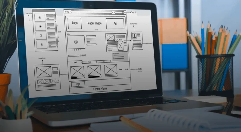 A laptop screen displaying a web design wireframe, positioned on a desk surrounded by office supplies such as pencils in a holder and a notebook.