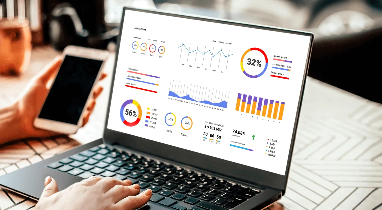 A person analyzing colorful data charts and graphs on a laptop screen, with a smartphone by their side on a busy office desk.