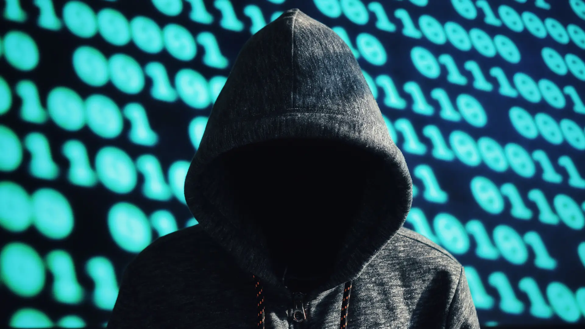Faceless hooded figure against a wall of glowing binary code, representing deepfake identity fraud