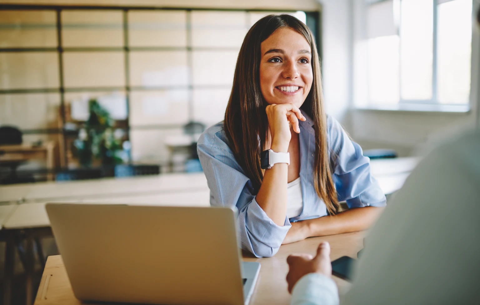 Smiling young woman in a casual blouse sits at a desk with a laptop, engaged in a conversation or interview in a modern, bright office setting.