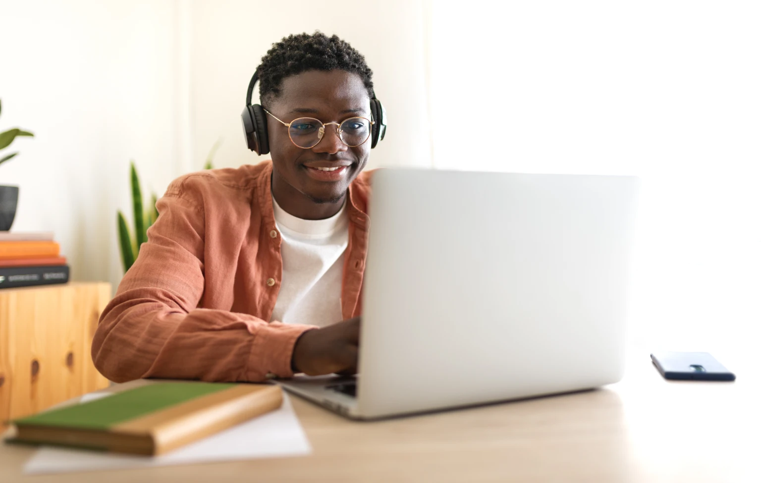 Smiling young man wearing headphones and glasses works on a laptop at a desk, with books and a smartphone nearby.