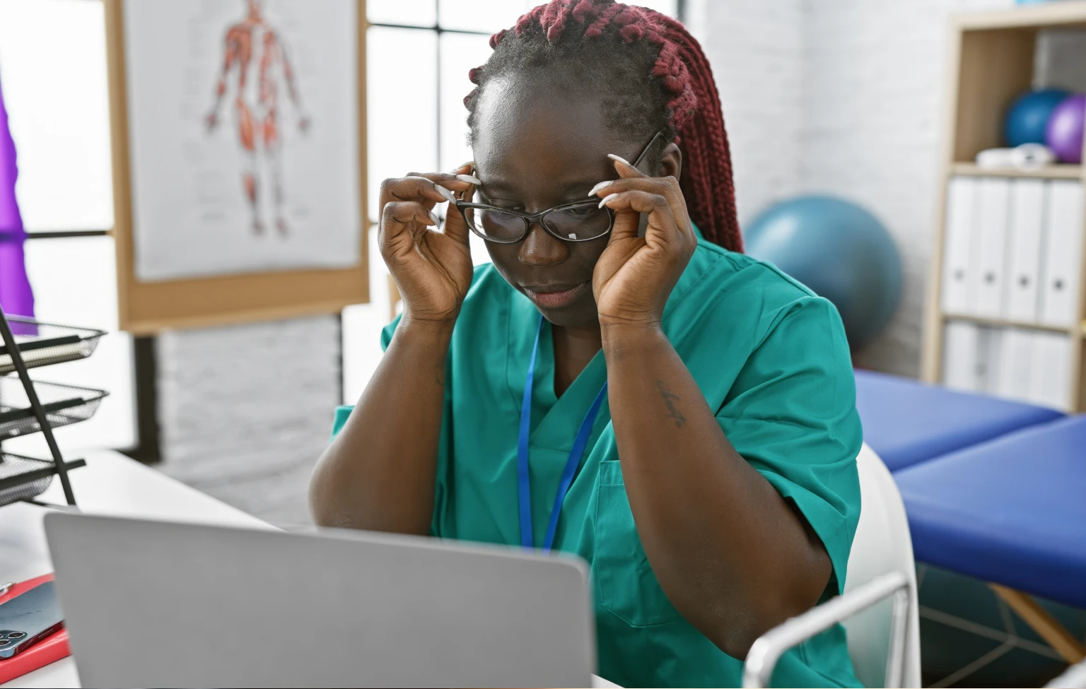 Occupational therapy student in scrubs adjusting glasses while studying on a laptop in a classroom or lab setting.