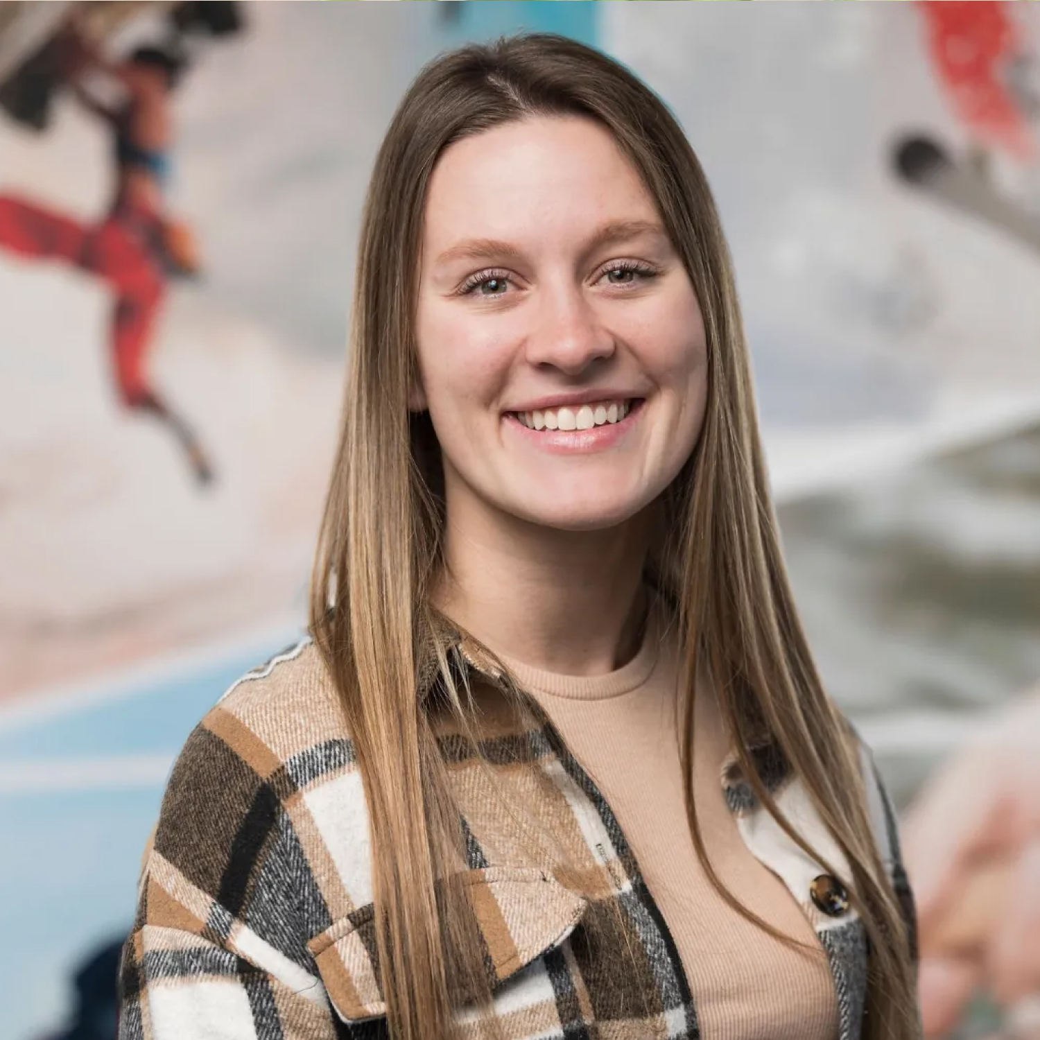 Kirstin Borge, OTD, smiling and standing in front of a blurred background featuring colorful athletic imagery. She has long straight hair and is wearing a tan shirt with a brown plaid jacket.