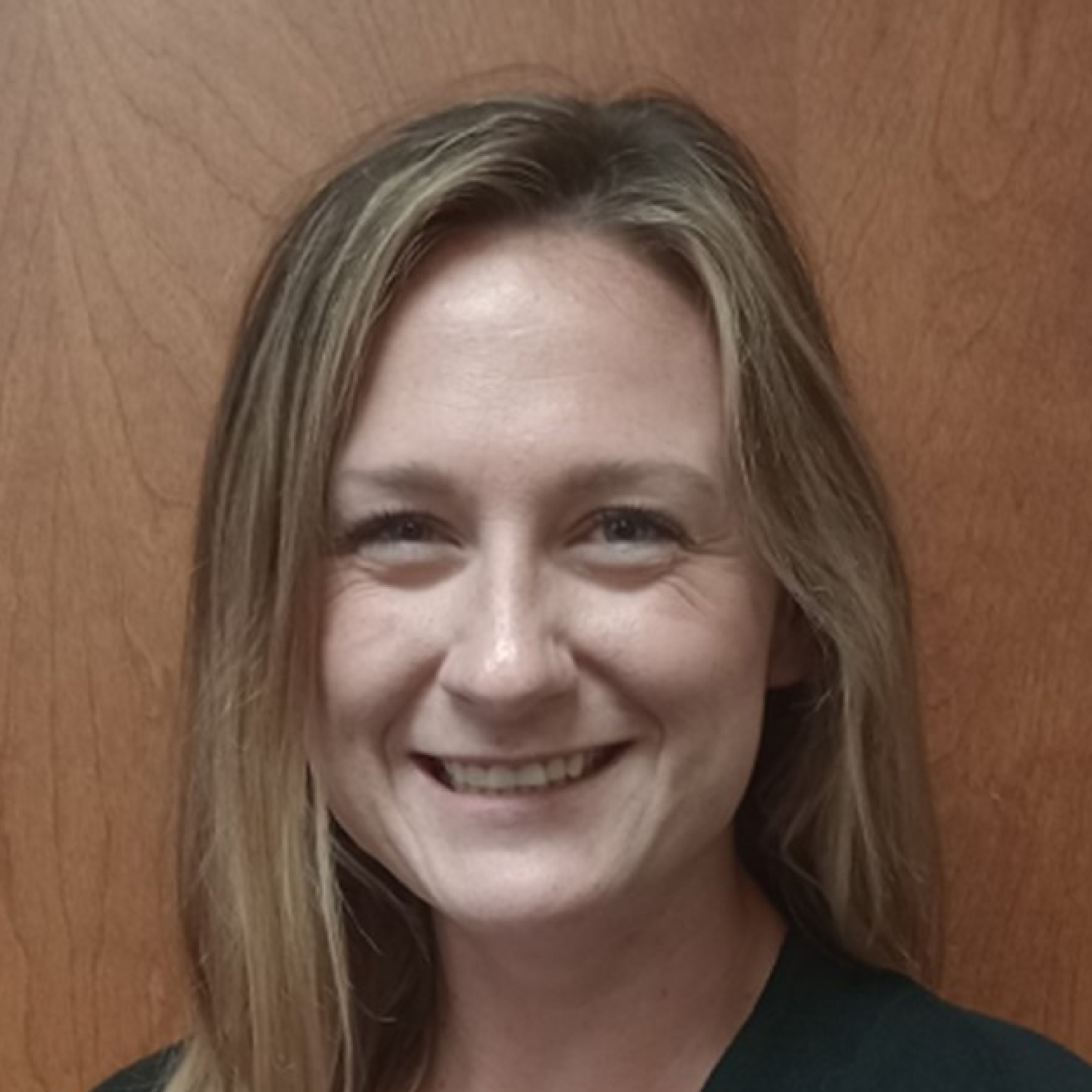 Kaitlin Burke smiling in front of a wooden background, wearing a dark top, with straight light brown hair parted slightly to the side.