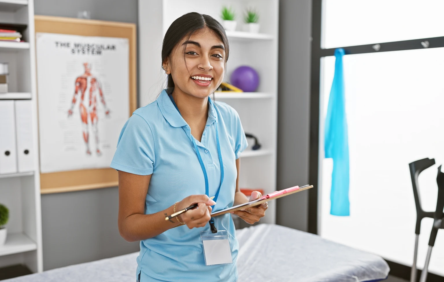 Occupational therapy student holding a clipboard in a clinical lab, standing near an exam table with anatomy charts and therapy equipment in the background.