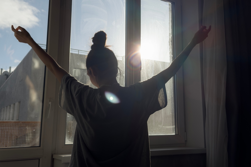 Person stretching and opening curtains to let sunlight in, symbolizing breaking out of the freeze response with gentle trauma tools