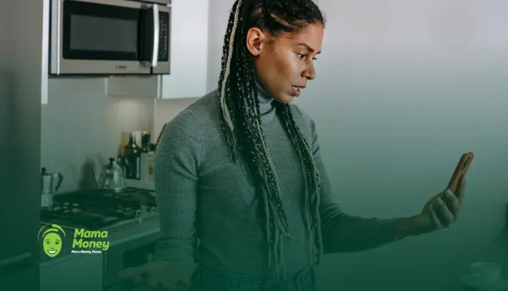 Concerned woman looking at her smartphone in a kitchen, representing awareness of online money transfer scams and fraud prevention.