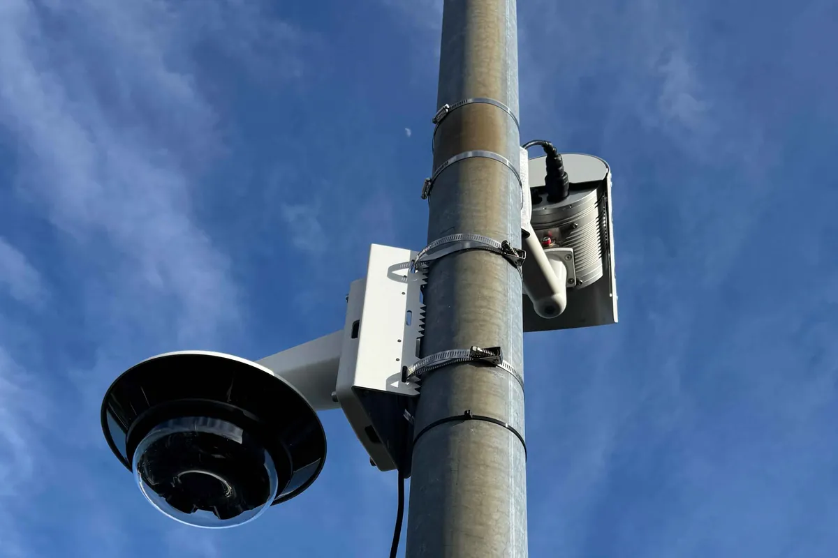 A multi-lens panoramic CCTV camera and a second camera unit mounted on a lighting column against a blue sky.