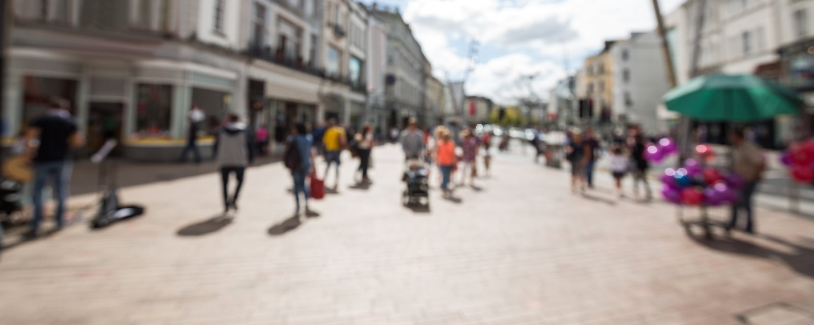 A busy pedestrianised town-centre street seen from a distance, with shoppers walking between shopfronts; the image is blurred so no individual can be identified.