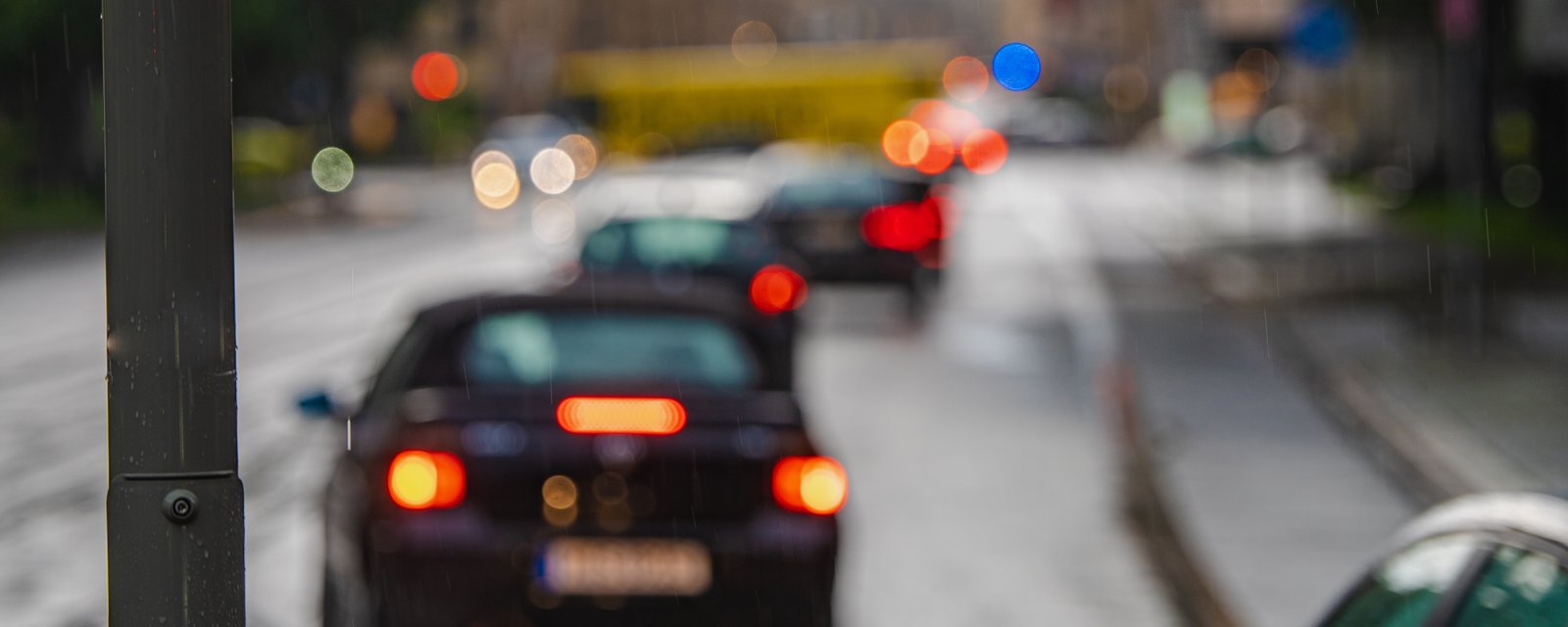Cars queuing on a wet city street at dusk, viewed from behind with brake lights showing, tram tracks running alongside the carriageway.