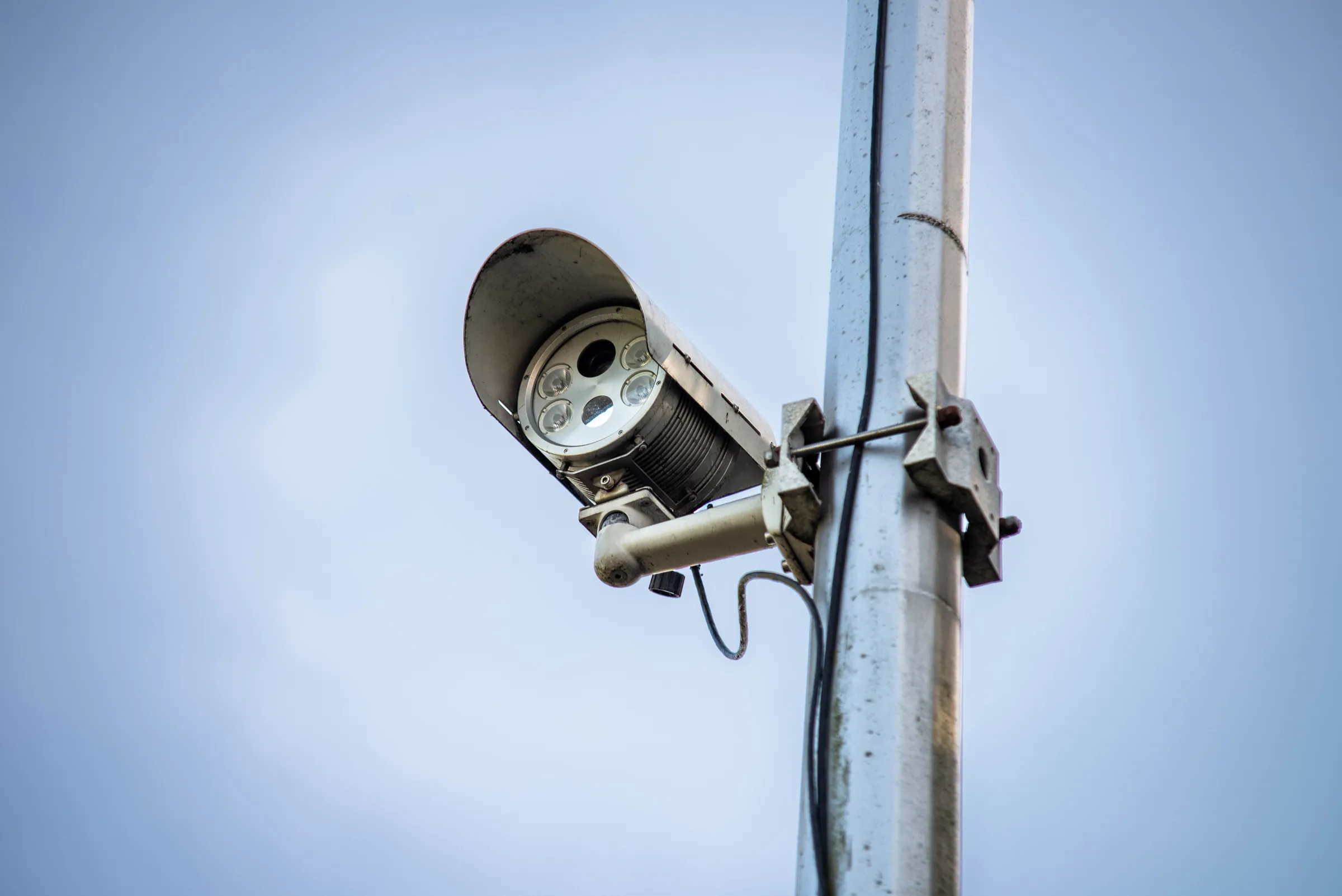 An ANPR camera with an infrared illuminator under a weather hood, mounted on a galvanised column against a pale sky.