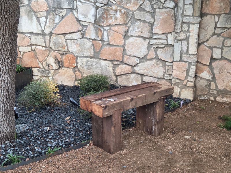 Handcrafted reclaimed wood garden bench positioned along a decomposed granite pathway beside custom steel planters and limestone wall landscaping.