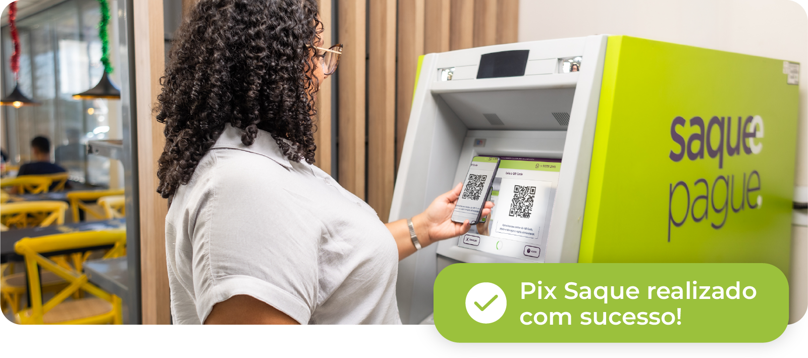 Mulher usando avental verde, de braços cruzados, em uma loja com potes de mantimentos no primeiro plano. Ao fundo, um caixa eletrônico ‘Saque e Pague’ está visível no lado direito.