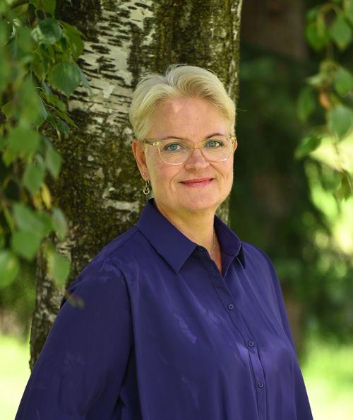 Livssynsåpen seremoniholder og terapeut Linda Rønold lener seg mot et bjørketre og ser i kamera. Hun har på seg en blå bluse.