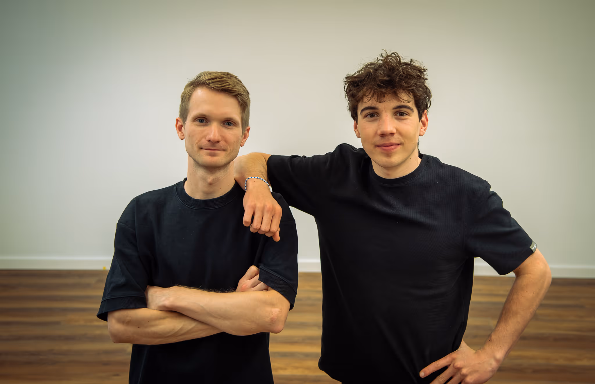 Zwei junge Männer in schwarzen T-Shirts stehen vor einer weißen Wand, einer legt seinen Arm auf die Schulter des anderen.