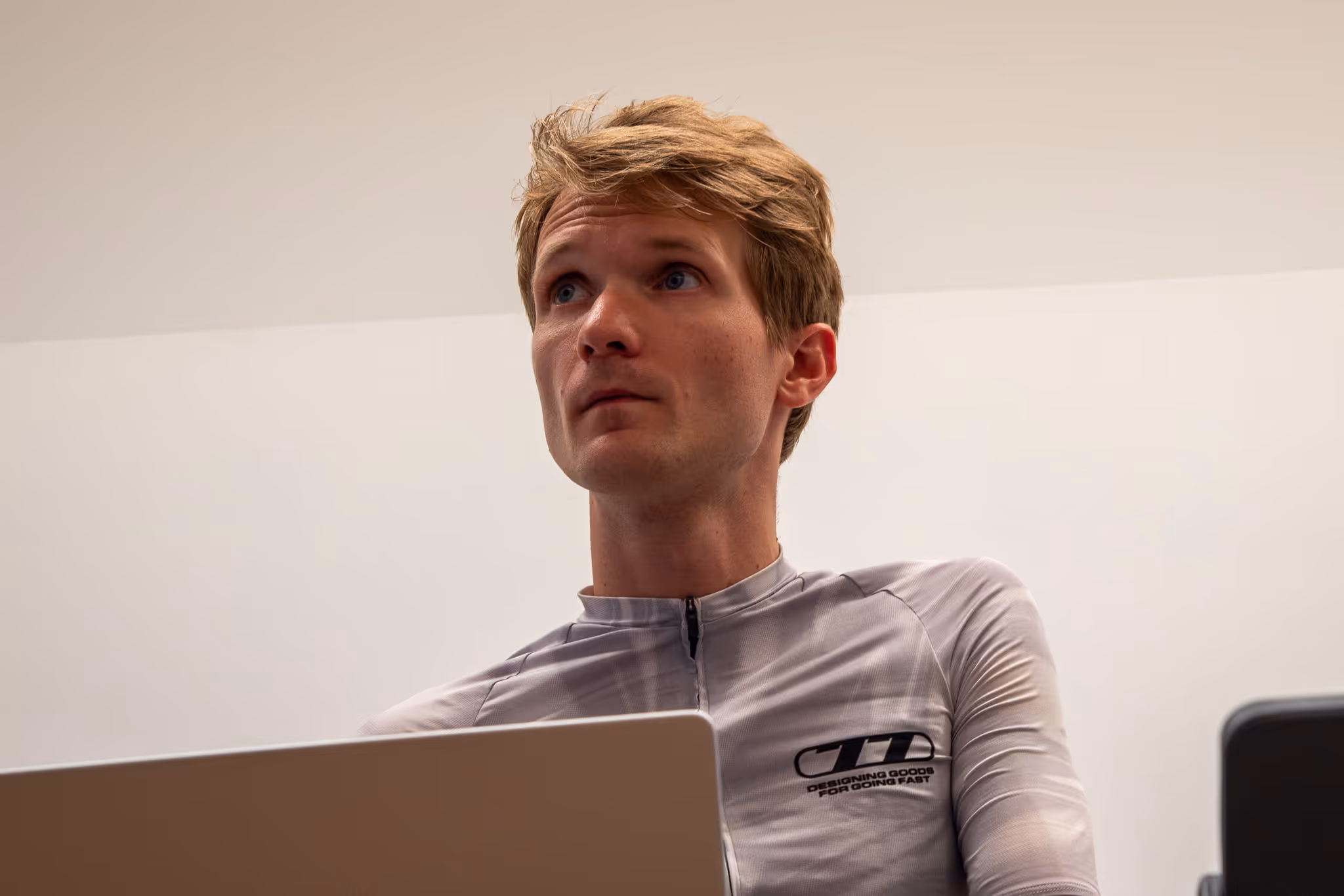 Junger Mann mit sportlichem Shirt sitzt vor einem Laptop und schaut nach oben.