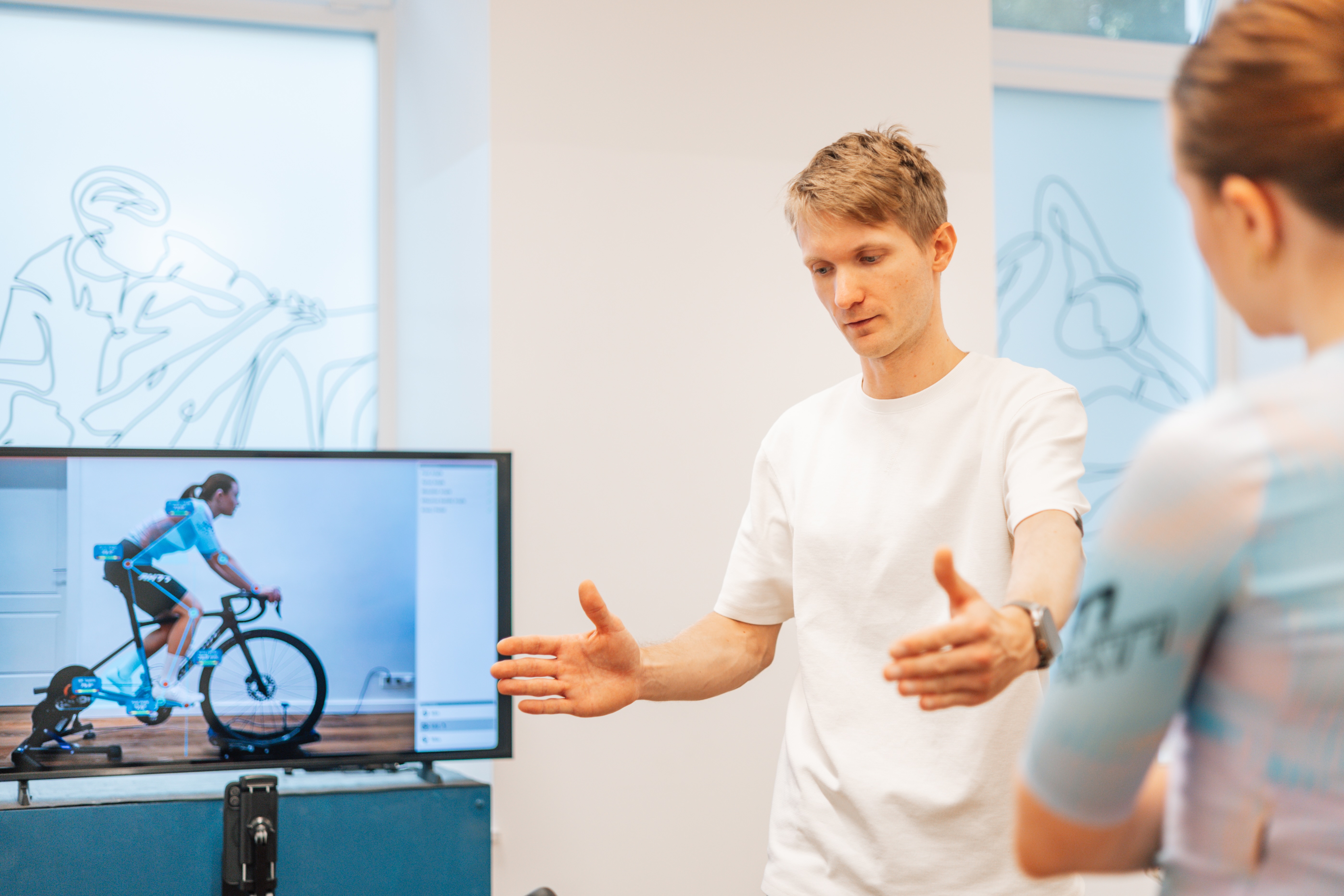 Trainer erklärt einer Radfahrerin die richtige Haltung beim Fahrradfahren anhand einer Analyse auf einem Bildschirm, der die Position eines Fahrers auf einem Fahrradergometer zeigt.