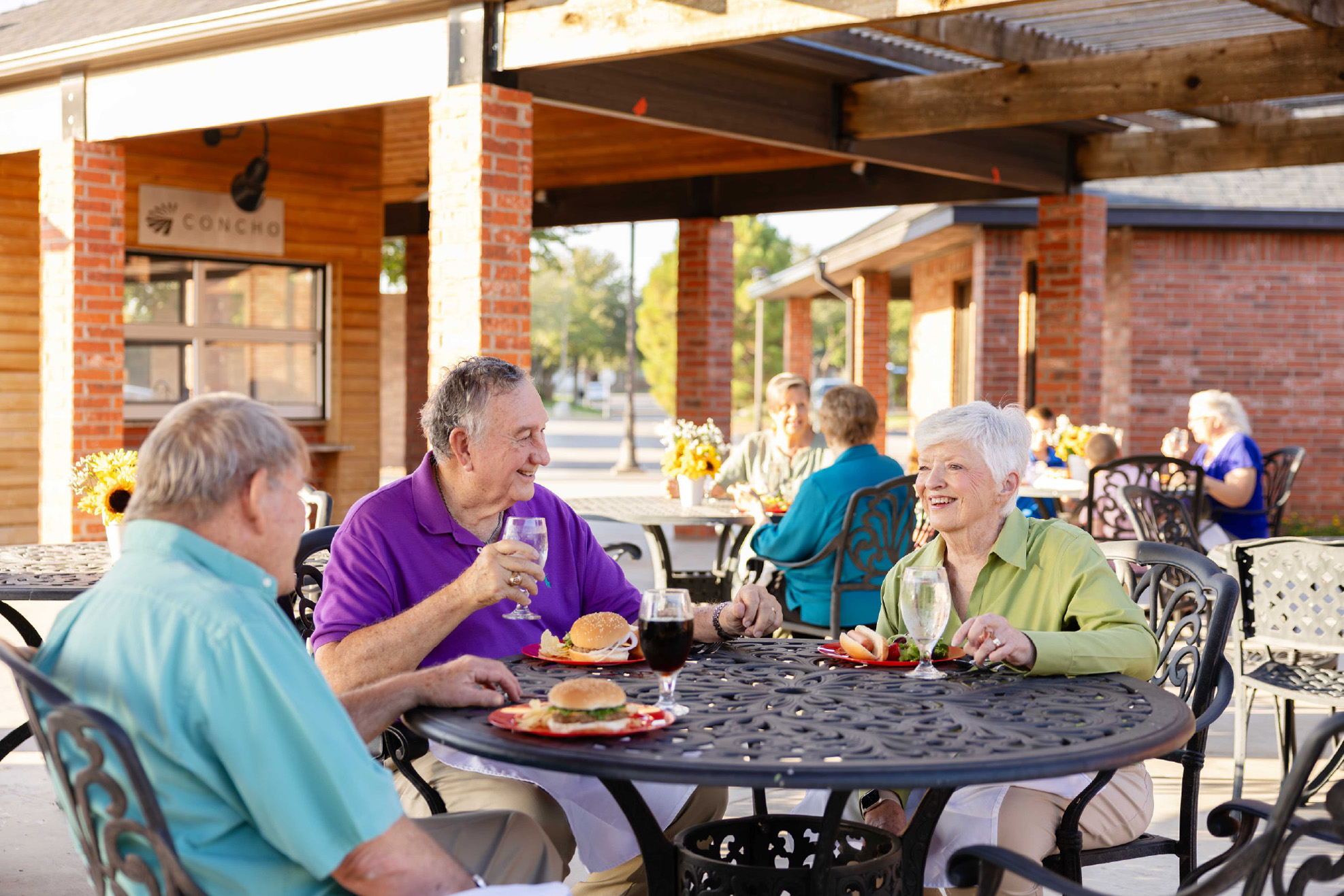 Three older adults sit at an outdoor table, enjoying food and drinks together.