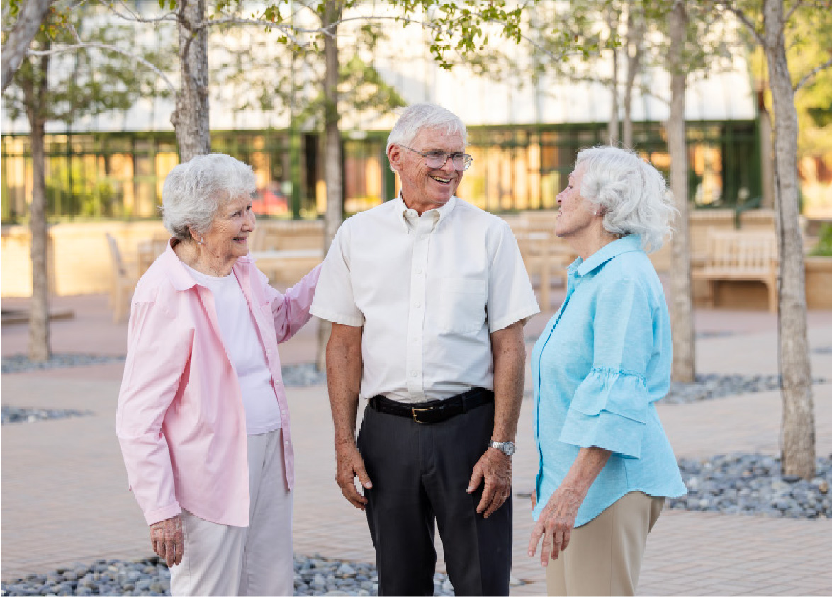 Three elderly people are standing together outdoors, smiling and talking.