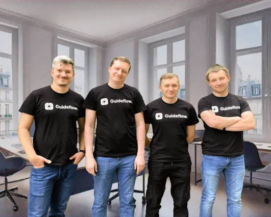 Four men standing side by side in an office, all wearing black Guideflow t-shirts, with windows and office chairs in the background.
