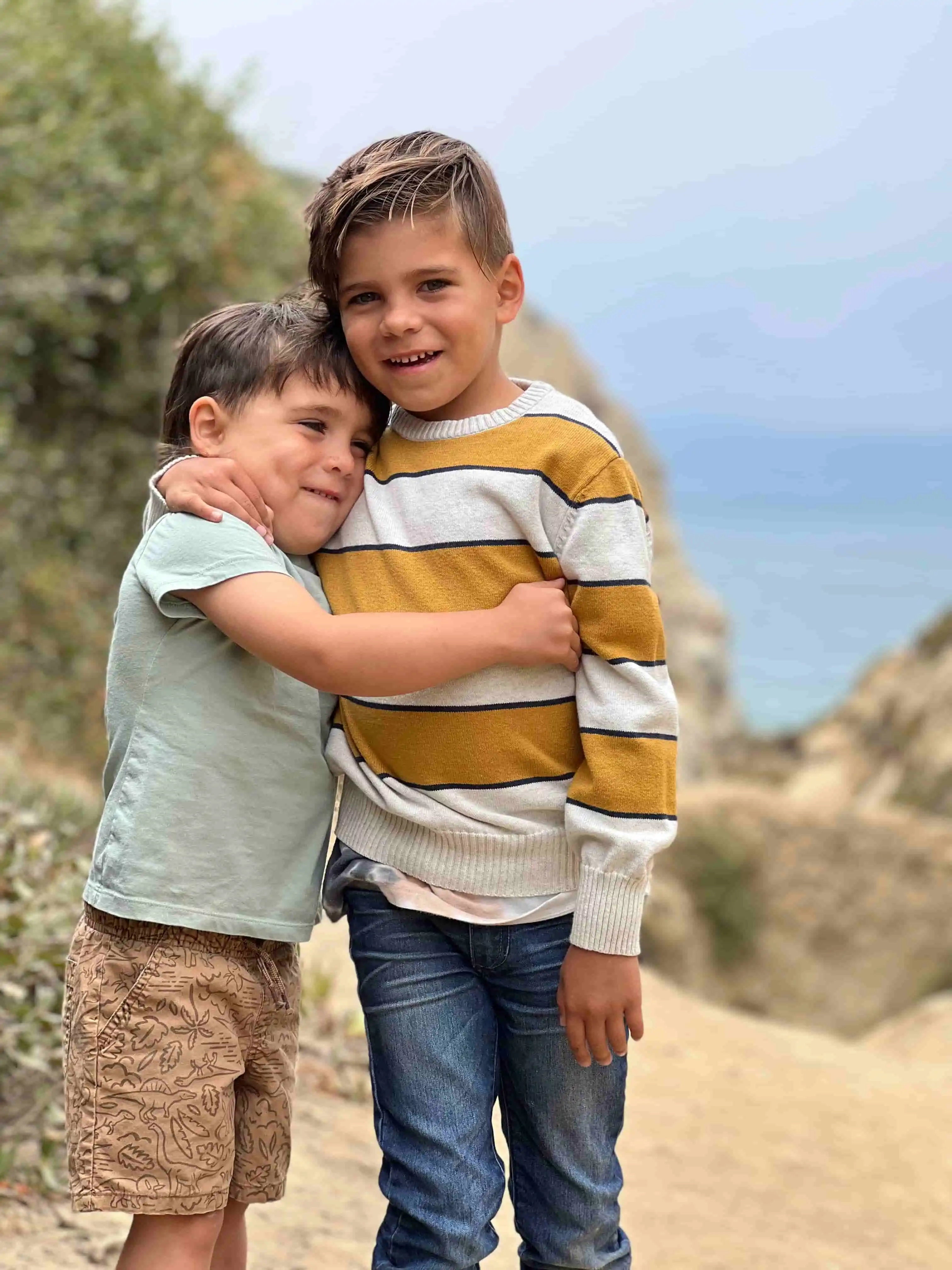 Two young brothers share a heartfelt hug along a coastal path, surrounded by soft cliffs and ocean air. Their natural smiles capture the essence of family connection and storytelling.