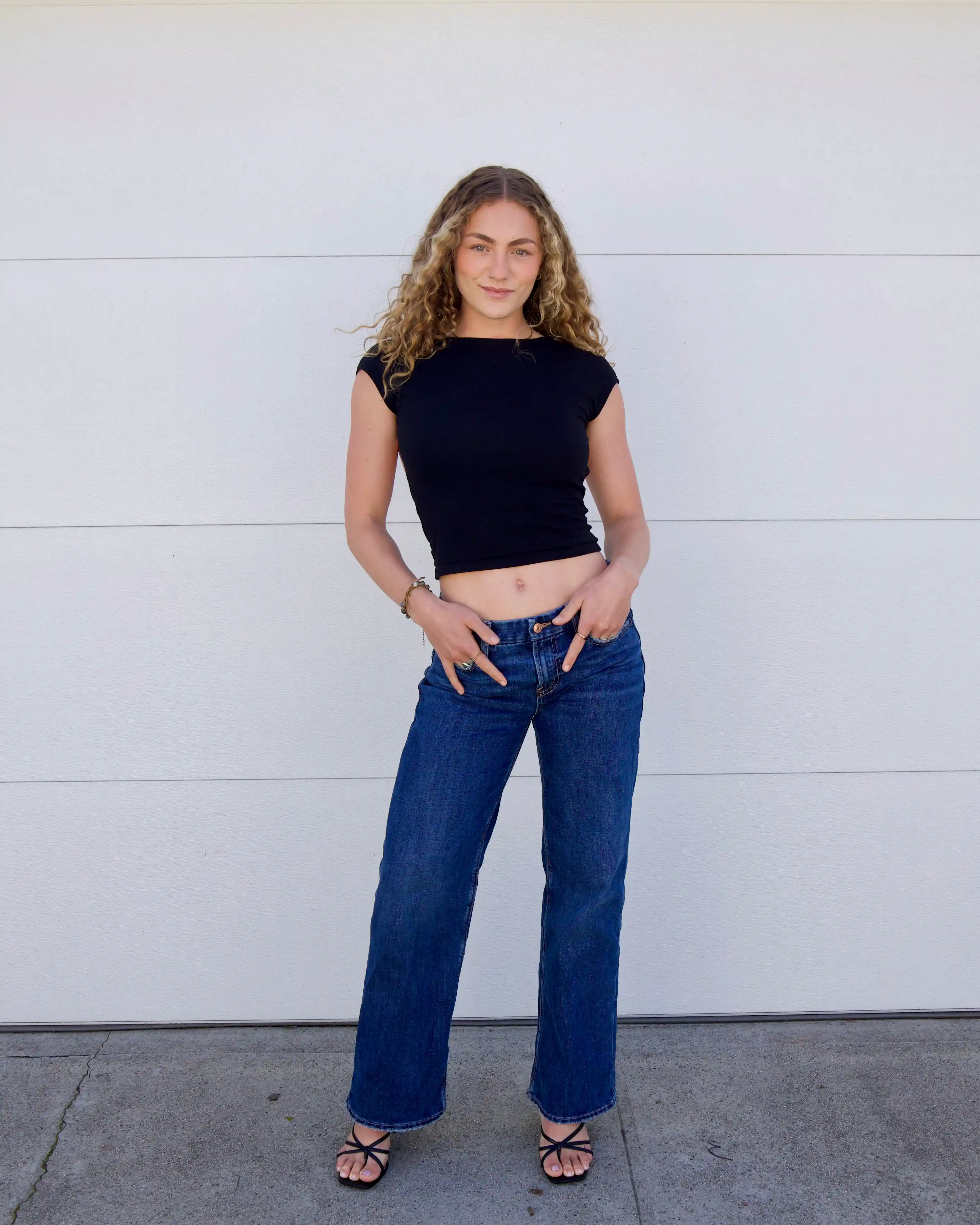 Model with blonde hair and green eyes posing with hands in her front pocket, wearing a fitted black t-shirt, dark blue straight-leg jeans, and strappy black kitten heels.