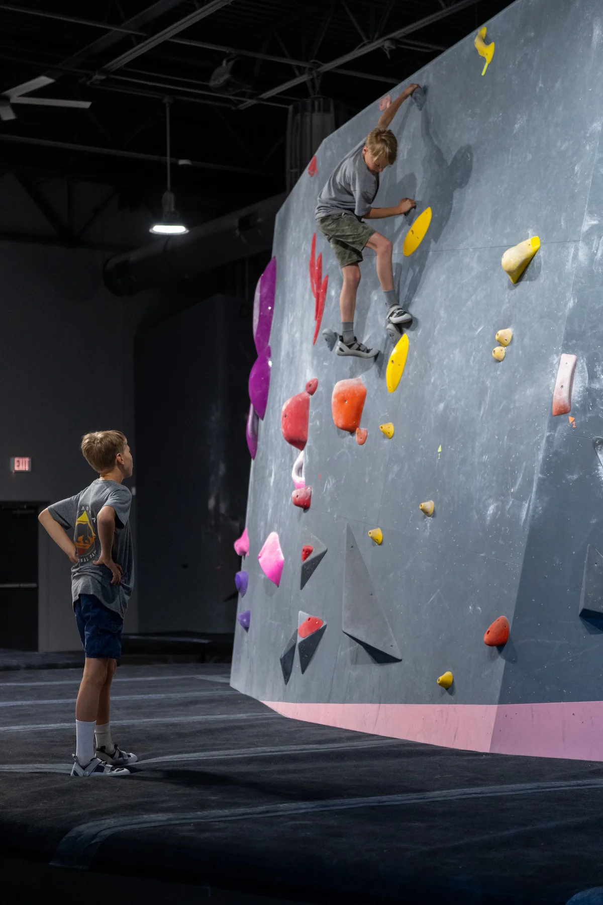 Young climber topping out a bouldering wall while a friend spots from below at Shift Climbing