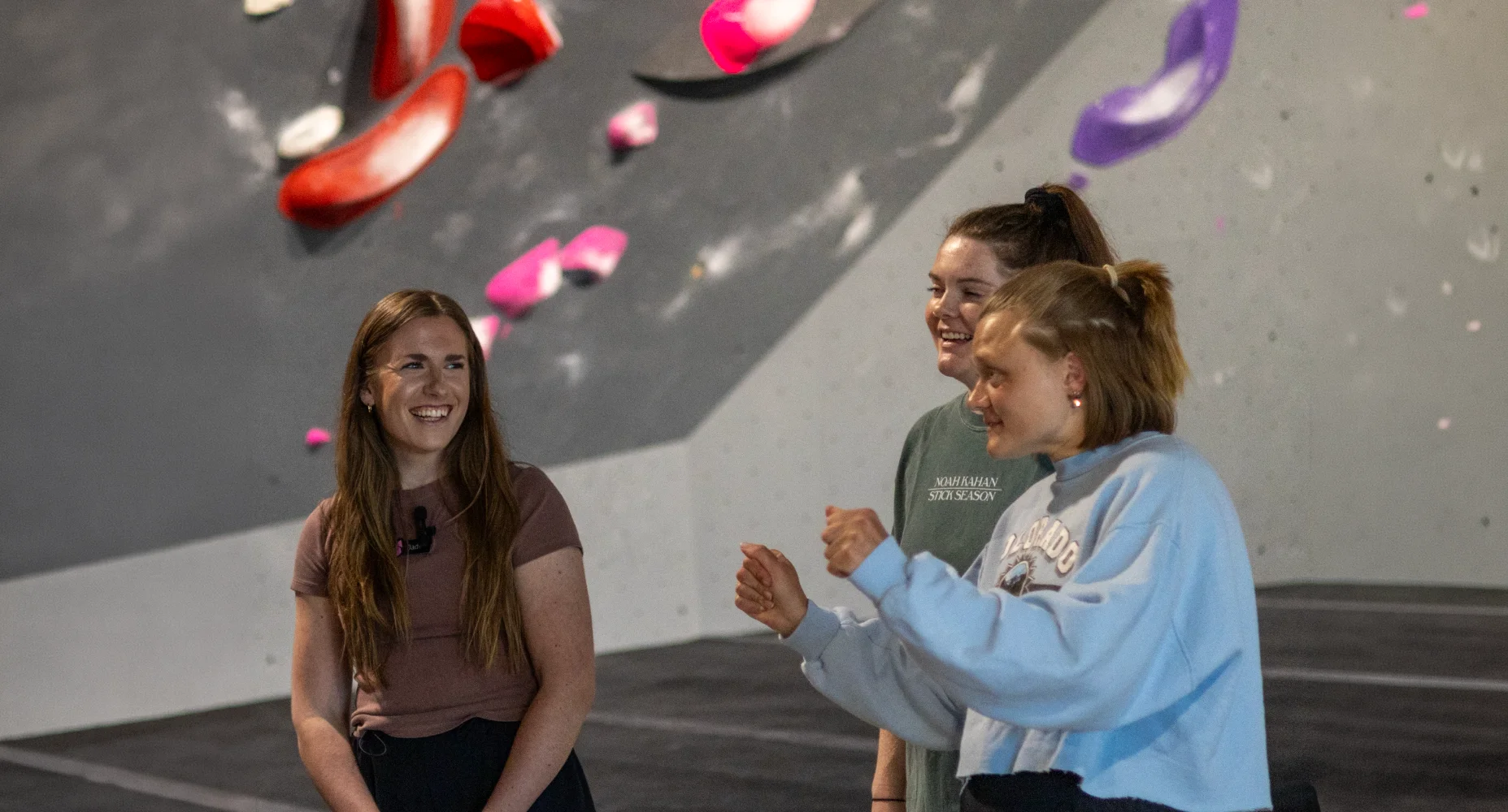Three women smiling and talking together on the mats during a private climbing instruction class at Shift Climbing gym.