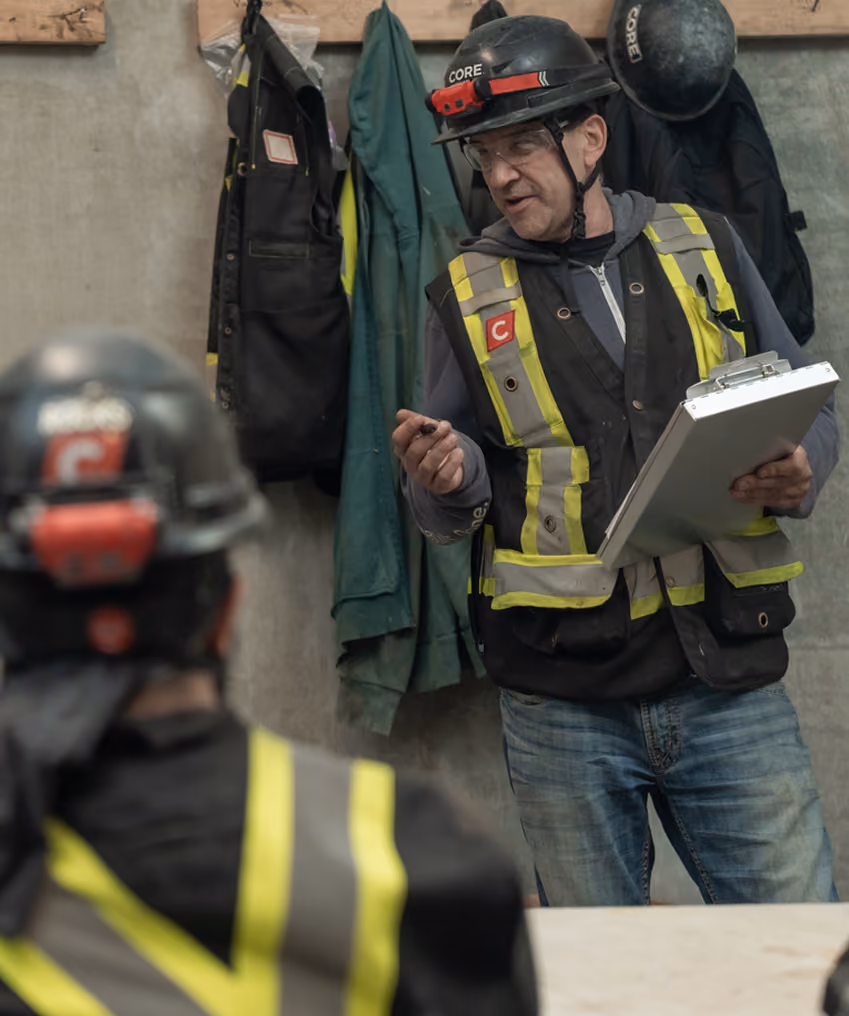Worker wearing a helmet and safety vest holding a clipboard, speaking to a colleague at a worksite.