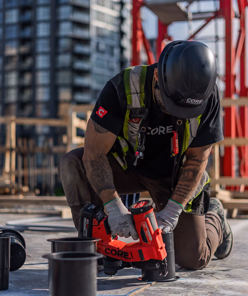 Construction worker kneeling and using a red CORE mechanical tool on a building site floor.