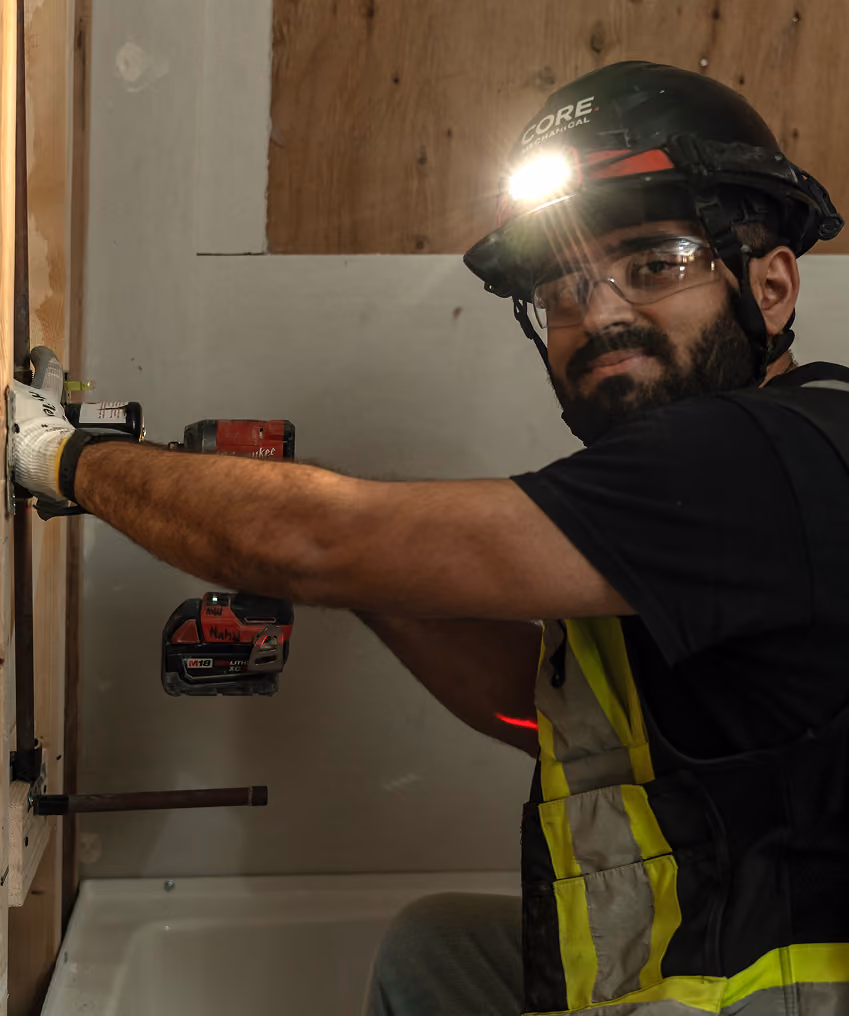Construction worker wearing hard hat with light using power drill on a wall indoors.